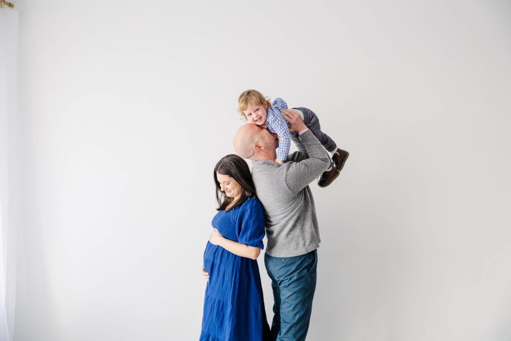 Family of four with dad holding son in the air and mom holding her belly in The White Room studio in West Knoxville