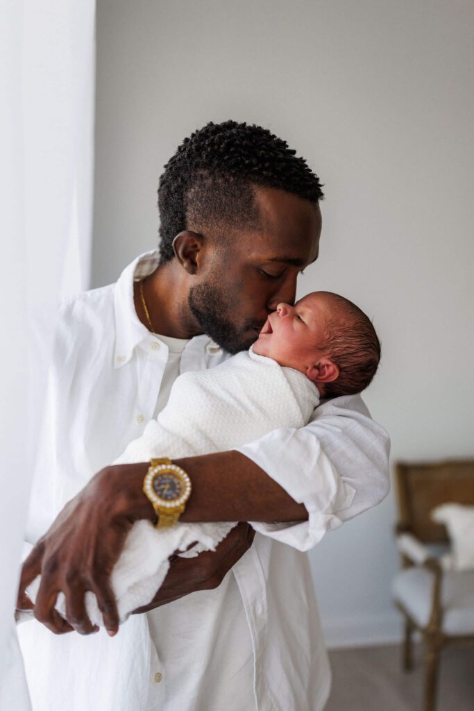 dad kissing newborn on cheek beside the windows