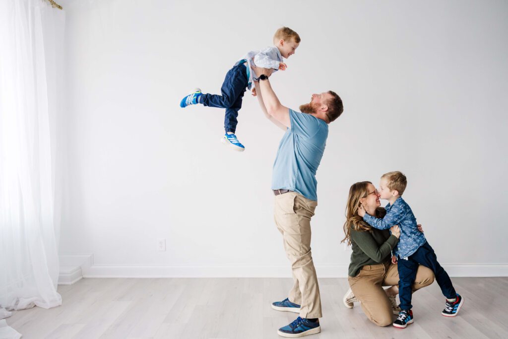 dad lifting son in the air in the white room studio while mom plays with other son behind him