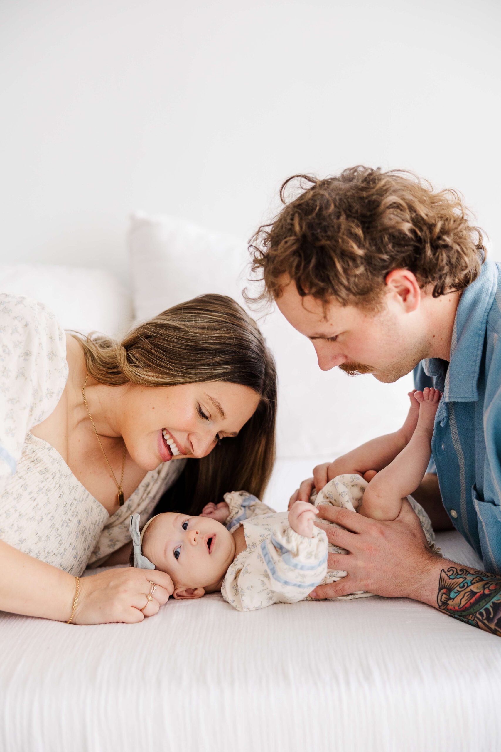 baby laying on bed with mom smiling over her and dad gazing at them both